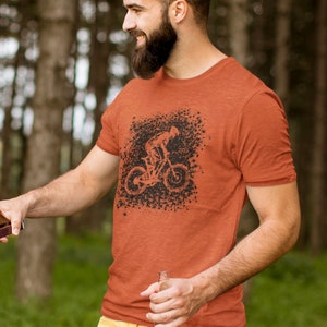 May include: A rust-coloured t-shirt with a black graphic of a person riding a mountain bike. The graphic is surrounded by a black splatter pattern.