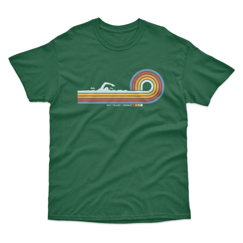 May include: A green t-shirt with a retro-style graphic of a swimmer in a wave. The graphic is in shades of orange, yellow, blue, and green. The text "EAT SLEEP SWIM" is printed below the graphic.