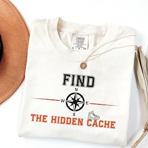 以下が含まれることがあります： クリーム色のTシャツで、黒いコンパスのグラフィック、黒い文字で「FIND」、オレンジ色の文字で「THE HIDDEN CACHE」とプリントされています。テキストの横には小さなオレンジ色のハイキングブーツのアイコンがあります。襟元にはComfort Colorsのブランドラベルが付いています。