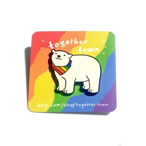 May include: An enamel pin featuring a white polar bear wearing a rainbow bandana. The pin is on a card with the text "together town" and "etsy.com/shop/togethertown".