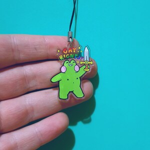 May include: A green frog holding a sword with a rainbow colored "Gay Rights" banner above its head. The frog is holding the sword in its right hand and has a pink blush on its cheeks. The frog is on a clear acrylic keychain.