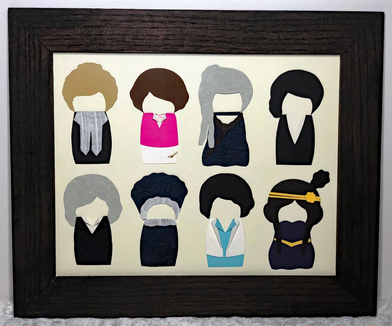 May include: A framed artwork featuring eight paper cutouts of people with different hairstyles and clothing. The artwork is set against a cream background and framed in a dark brown frame.
