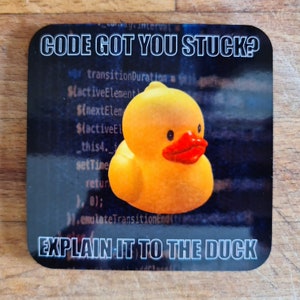 以下が含まれることがあります： 黒い背景にコードのテキストと「CODE GOT YOU STUCK?」と「EXPLAIN IT TO THE DUCK」の文字が書かれた、赤いクチバシの黄色いゴム製のオモチャの鴨。
