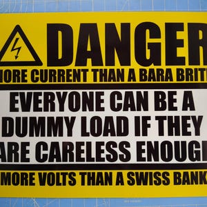 Könnte beinhalten: Ein rechteckiges gelb-weißes Schild mit schwarzem Text und einem Blitzsymbol. Das Schild trägt die Aufschrift: DANGER, MORE CURRENT THAN A BARA BRITH, EVERYONE CAN BE A DUMMY LOAD IF THEY ARE CARELESS ENOUGH, MORE VOLTS THAN A SWISS BANK.