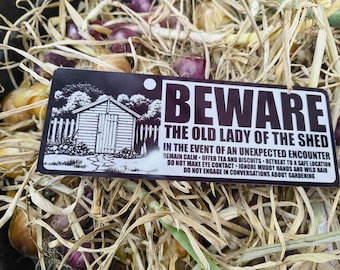 May include: A rectangular black and white sign featuring a graphic of a shed and the text: BEWARE THE OLD LADY OF THE SHED. IN THE EVENT OF AN UNEXPECTED ENCOUNTER REMAIN CALM - OFFER TEA AND BISCUITS - RETREAT TO A SAFE LOCATION DO NOT MAKE EYE CONTACT - IGNORE MUDDY HANDS AND WILD HAIR DO NOT ENGAGE IN CONVERSATIONS ABOUT GARDENING.