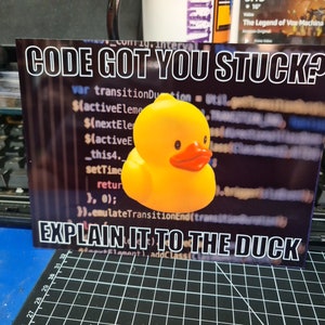 May include: A yellow rubber duck with a red beak and eyes is in front of a computer screen with code on it. The text on the image says "CODE GOT YOU STUCK? EXPLAIN IT TO THE DUCK".