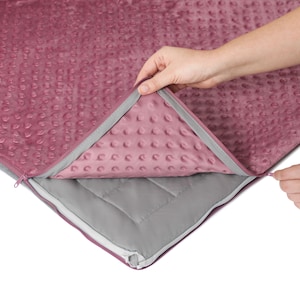 May include: A pink weighted blanket with a zipper. The blanket is made of soft, plush fabric with a subtle pattern. The zipper allows the blanket to be opened and the inner layers can be seen.