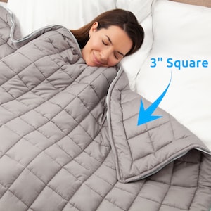May include: A gray weighted blanket with a zipper closure. The blanket is quilted with 3-inch square compartments. A person is sleeping under the blanket.