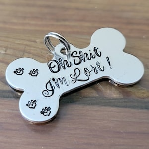May include: A silver-colored, bone-shaped pet tag with the phrase "Oh Shit I'm Lost!" in black script. The tag also features three paw print designs. A small metal ring is attached for easy attachment to a pet's collar.