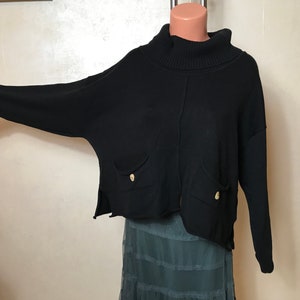 May include: Black sweater with a turtleneck collar and long sleeves. The sweater has two front pockets with gold-colored buttons. The bottom hem is uneven. The sweater is displayed on a mannequin.