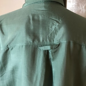 Op de afbeelding: Een zijden knoopsluiting overhemd in saliegroen met een enkele knoop op de achterkant van de kraag.