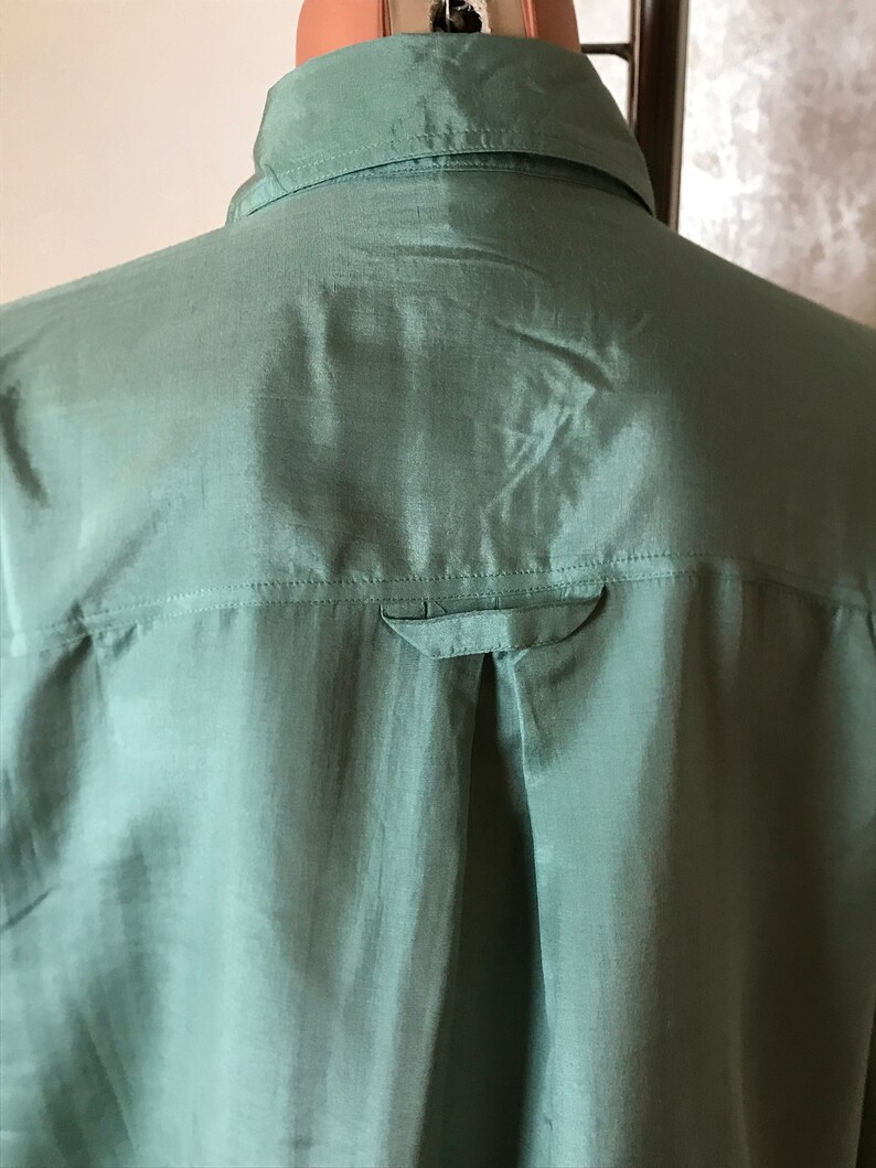 Op de afbeelding: Een zijden knoopsluiting overhemd in saliegroen met een enkele knoop op de achterkant van de kraag.