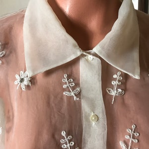 May include: A white sheer blouse with a collared neckline and button closure. The blouse is decorated with embroidered floral designs in white beads.