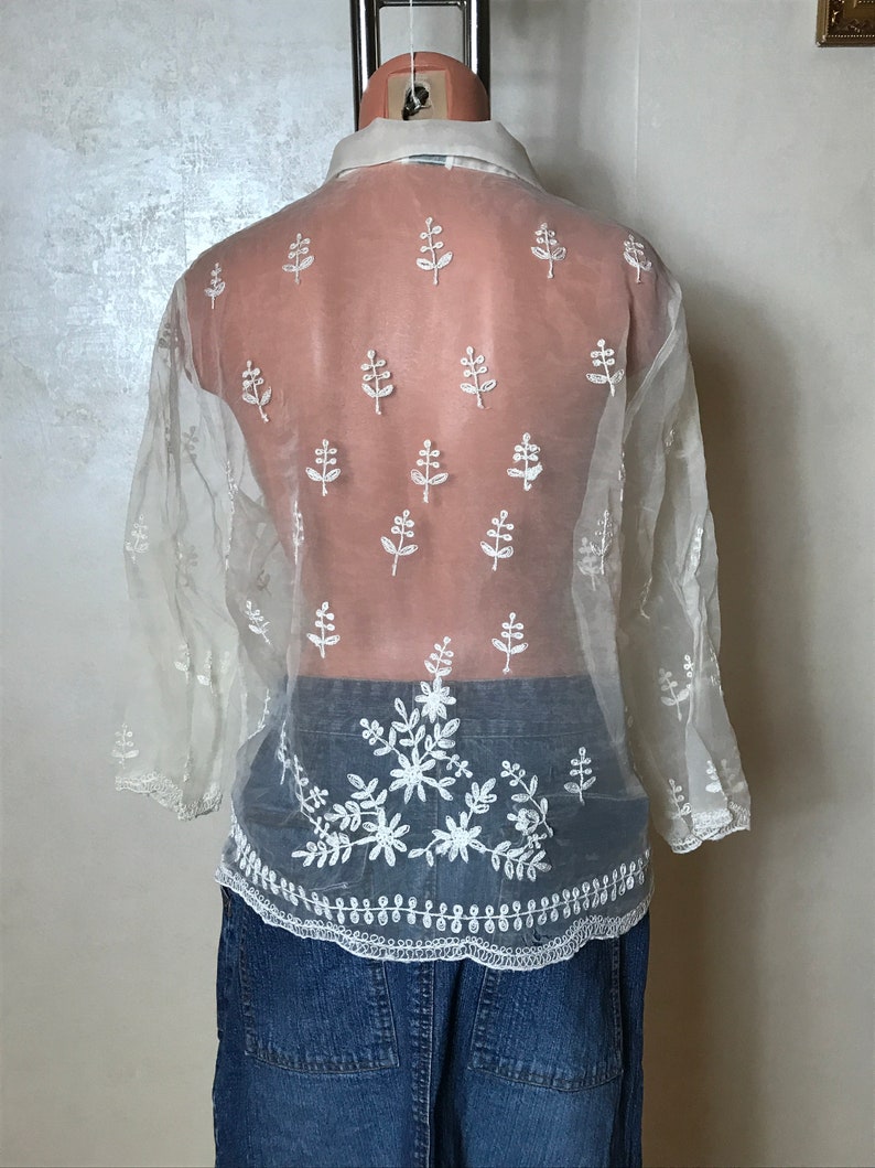 May include: A white sheer blouse with embroidered floral details. The blouse has a collared neckline and long sleeves. The embroidery is in a delicate floral pattern.