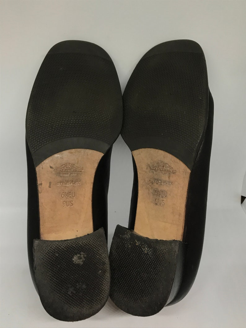 May include: Black leather shoes with a low heel. The soles are black rubber with a textured pattern. The shoes are labelled "6 1/2 EU 9 US".