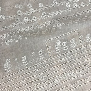 May include: White cotton fabric with a delicate embroidered pattern of flowers and leaves. The fabric has a subtle sheen and a soft texture.