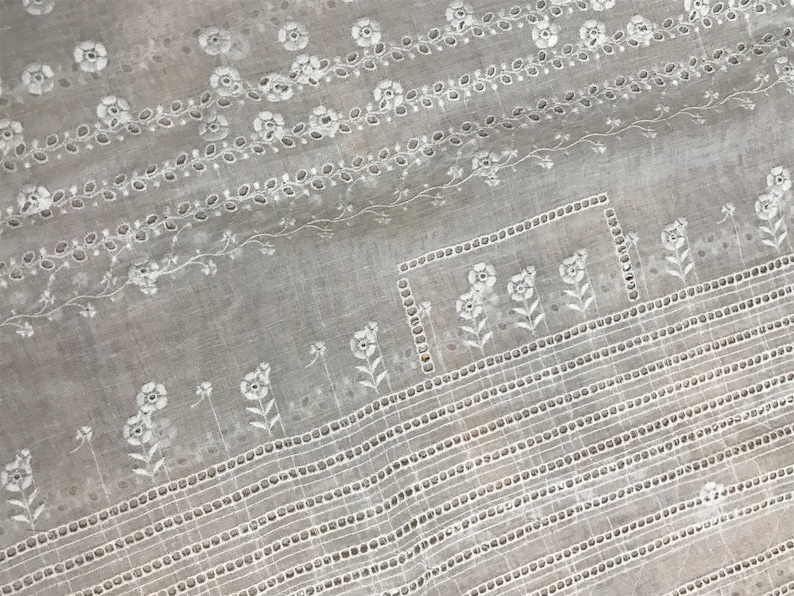 May include: White cotton fabric with a delicate embroidered pattern of flowers and leaves. The fabric has a subtle sheen and a soft texture.