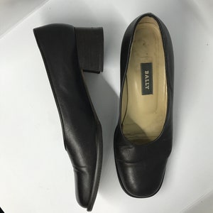 May include: A pair of black leather heeled shoes with a square toe. The shoes have a low block heel and the brand name "BALLY" is visible on the inside of one shoe.