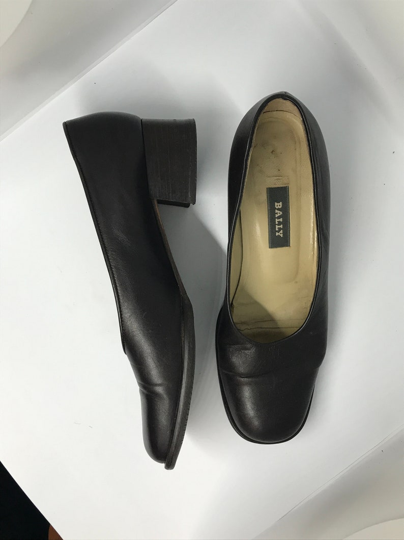 May include: A pair of black leather heeled shoes with a square toe. The shoes have a low block heel and the brand name "BALLY" is visible on the inside of one shoe.