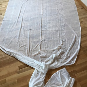 May include: A white sheer fabric with a delicate embroidered pattern. The fabric is folded and laid out on a wooden floor.