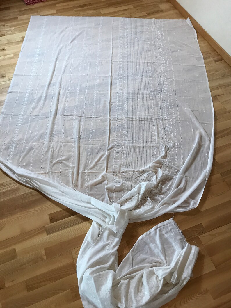 May include: A white sheer fabric with a delicate embroidered pattern. The fabric is folded and laid out on a wooden floor.