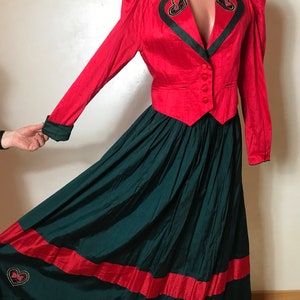 Op de afbeelding: Een tweedelige outfit in rood en groen, bestaande uit een rood jasje en een groene rok. Het jasje heeft een V-hals en een enkele knoopsluiting. De rok is lang en heeft een rode band aan de zoom. Beide delen zijn versierd met geborduurde harten.