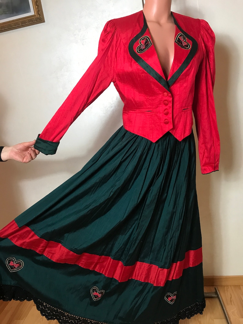 May include: A red and green two-piece outfit with a red jacket and a green skirt. The jacket has a V-neckline and a single button closure. The skirt is full-length and has a red band at the hem. Both pieces feature embroidered hearts.