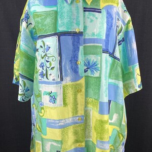 May include: A short-sleeved button-down shirt with a colorful abstract pattern. The shirt features a patchwork design with squares of blue, green, and yellow, adorned with floral accents. The shirt is displayed on a white mannequin.