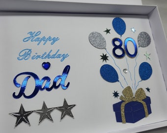 Può includere: Un biglietto bianco con decorazioni di glitter blu e argento. Il biglietto dice "Happy Birthday Dad" e ha una scatola regalo blu con un fiocco d'oro e il numero 80 in glitter blu.