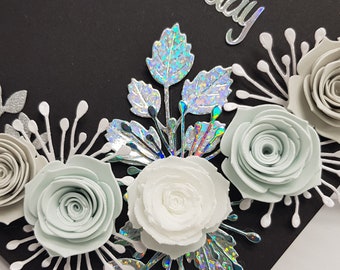 May include: A black card with a white and silver floral design. The flowers are made of paper and are in shades of white, gray, and light blue. The card has the word "Happy" written on it in silver glitter.