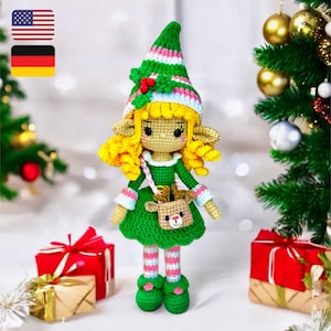Könnte beinhalten: Eine gehäkelte Elfenpuppe mit blonden Haaren, einem grünen Kleid und einem rosa-weiß gestreiften Rock. Der Elf hält ein kleines gehäkeltes Rentier in der Hand. Die Puppe steht vor einem Weihnachtsbaum und Geschenken.