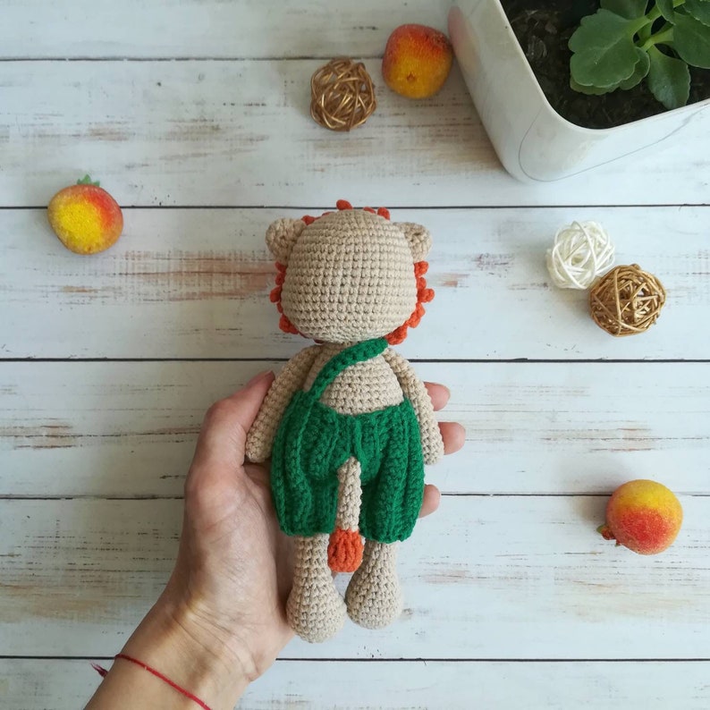 May include: A crocheted lion toy with a green and brown outfit. The toy is being held in a person's hand.