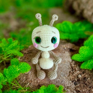 Pu&ograve; includere: Un giocattolo di formica amigurumi all'uncinetto con occhi verdi e guance rosa. La formica &egrave; seduta su una superficie marrone con vegetazione verde sullo sfondo.
