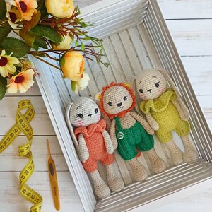 May include: Three crocheted stuffed animals in a white wooden box. The animals are a lion, a bunny, and a bear. The lion is wearing a green and white outfit, the bunny is wearing an orange and white outfit, and the bear is wearing a yellow and white outfit.