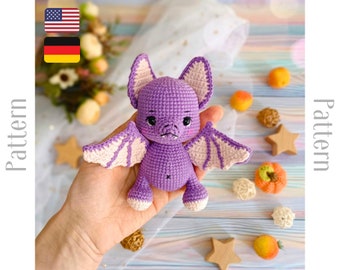 Puede incluir: Juguete de amigurumi de murciélago de crochet morado con alas rosas. El murciélago tiene una cara linda con ojos negros y una nariz pequeña. El juguete está sentado en una mano.
