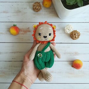 May include: A crocheted lion toy with a green and brown outfit. The lion has a brown face with orange hair and a green button on its chest.
