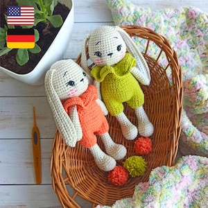 May include: Two crocheted bunny amigurumi toys in a wicker basket. One bunny is wearing an orange outfit and the other is wearing a green outfit. Both bunnies have white yarn for their ears and bodies.