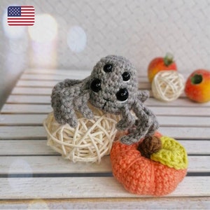 May include: A crocheted gray spider with black eyes sitting on a woven straw ball and a crocheted orange pumpkin with a green stem. The spider and pumpkin are on a white wooden surface.