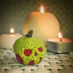 May include: A green crocheted apple with a red skull design. The apple is sitting on a white wicker surface with three candles in the background.