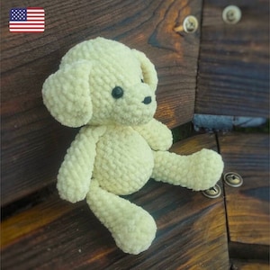 May include: A crocheted cream-colored plush dog toy sitting on a wooden surface. The dog has black eyes and a black nose.