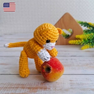 Puede incluir: Un juguete de gato de crochet naranja y blanco sosteniendo una fruta roja. El gato está sentado sobre una superficie de madera.