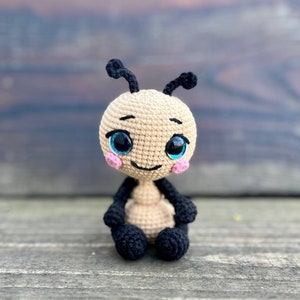 Pu&ograve; includere: Un giocattolo amigurumi all'uncinetto a forma di formica. La formica &egrave; beige con gambe e antenne nere. Ha grandi occhi blu e guance rosa.