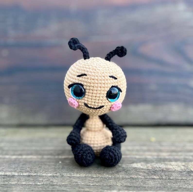 Pu&ograve; includere: Un giocattolo amigurumi all'uncinetto a forma di formica. La formica &egrave; beige con gambe e antenne nere. Ha grandi occhi blu e guance rosa.