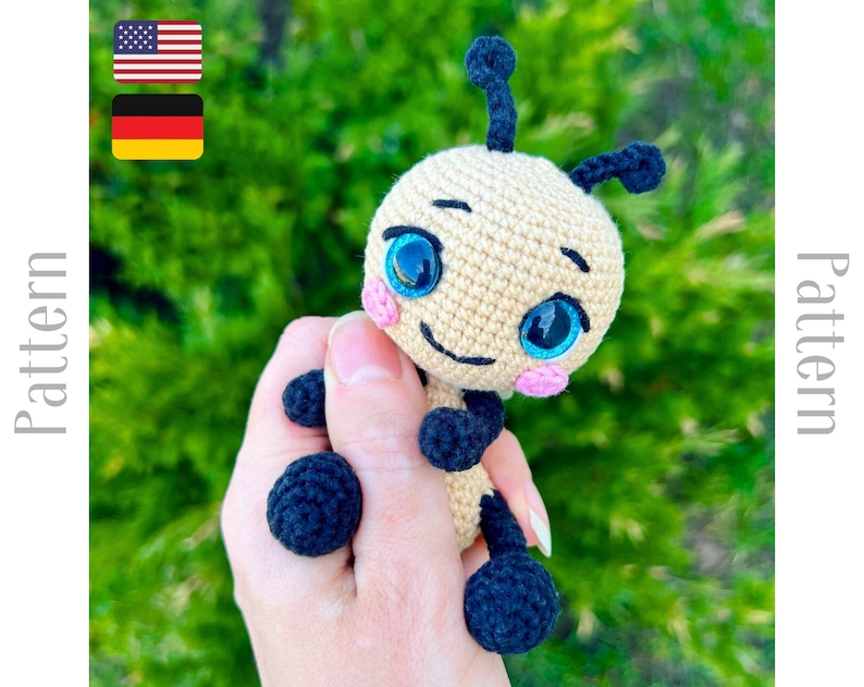 Pu&ograve; includere: Giocattolo ape amigurumi all'uncinetto in filato beige e nero, con grandi occhi blu e un naso rosa. L'ape &egrave; tenuta in una mano.