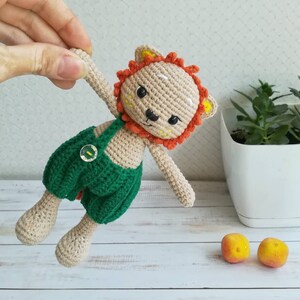 May include: A crocheted lion toy wearing a green and white overall with a button. The lion has orange and yellow mane and white facial features.