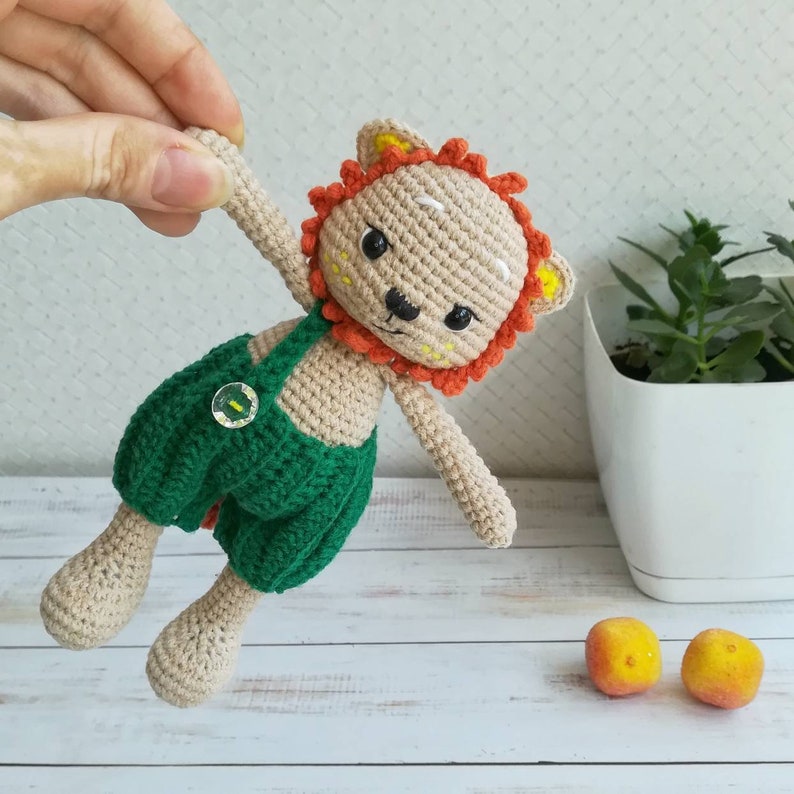 May include: A crocheted lion toy wearing a green and white overall with a button. The lion has orange and yellow mane and white facial features.