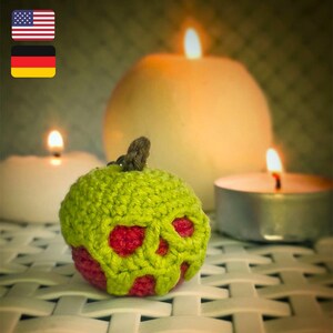 May include: A green crocheted apple with a red skull design. The apple is sitting on a white surface with two lit candles in the background.