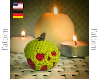 May include: A green crocheted apple with a red skull design. The apple is sitting on a white surface with two lit candles in the background.