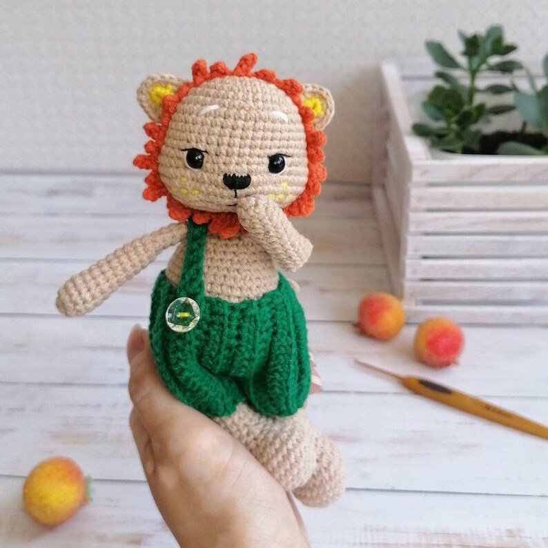 May include: A crocheted lion toy with orange mane, brown fur, green overalls, and a yellow button. The toy is being held by a hand.