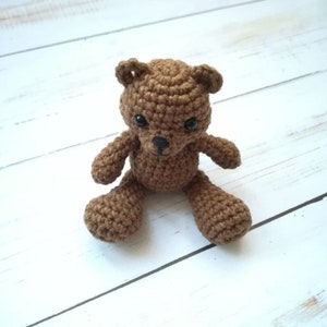 May include: A small brown crocheted teddy bear sitting on a white wooden surface. The bear has black eyes and a black nose.